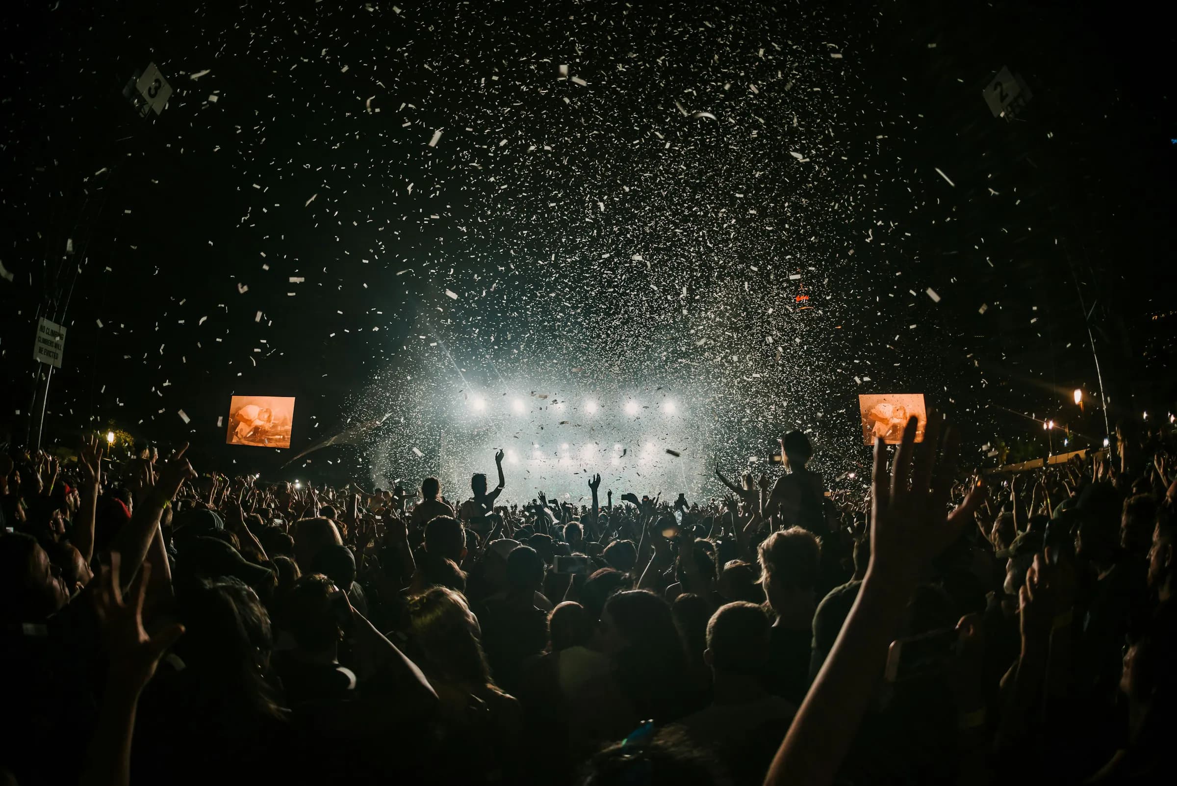 Confetti falling over a concert crowd in front of bright stage lights.